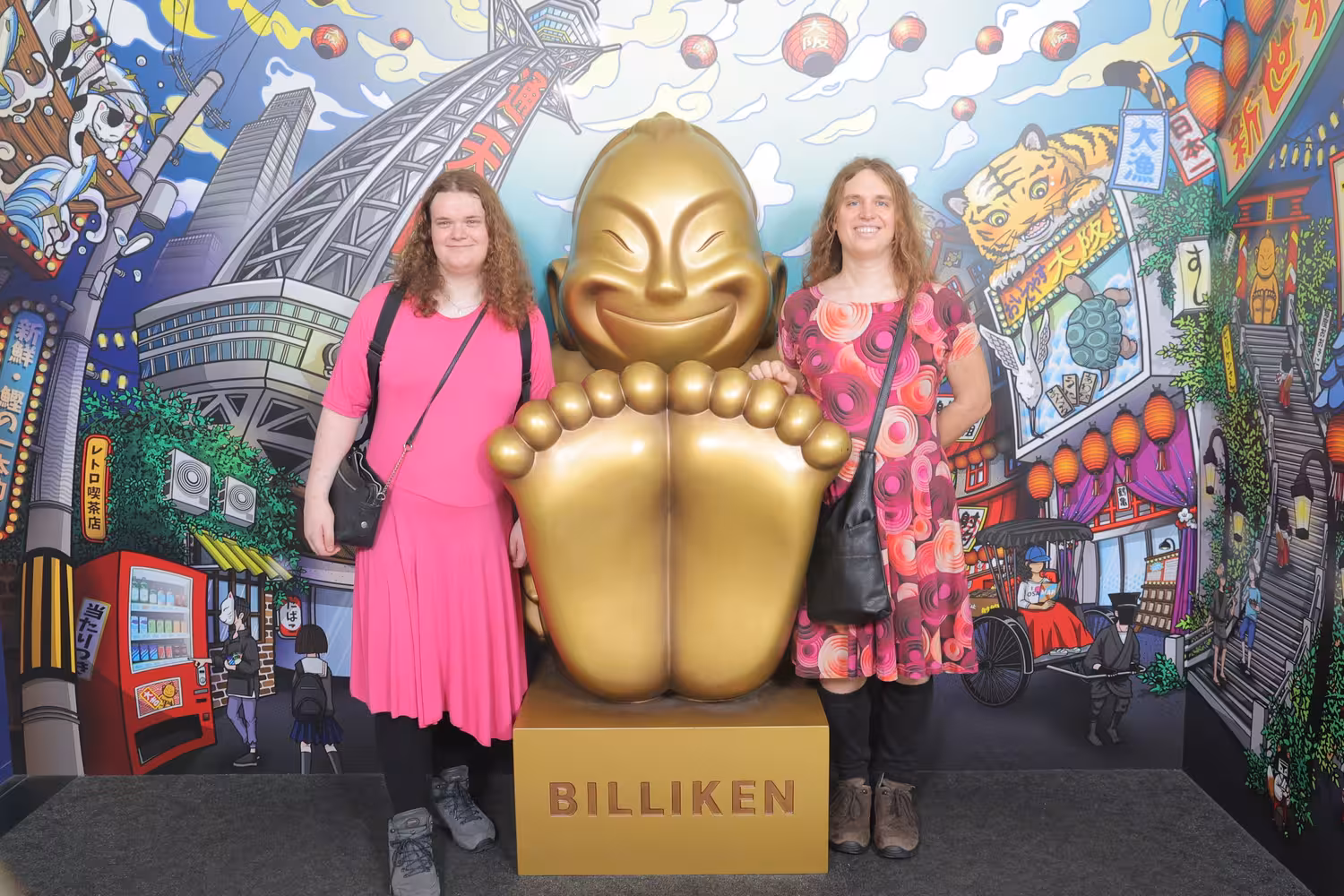Senna and Fiona standing on the sides of a golden statue with huge feet in front of a colorful modern style drawing of Osaka.