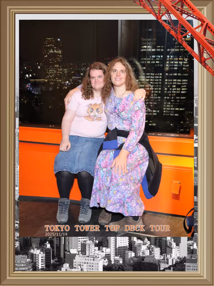 Senna and Fiona sitting on a side-rail embracing each other in fron of a window showing the city with a decorative digital frame.