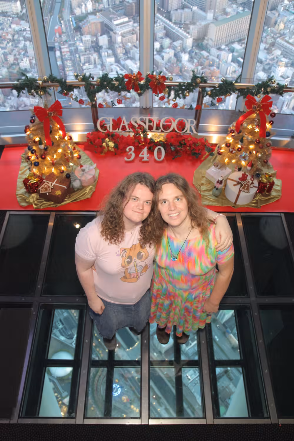 The official picture for them same place. This time Senna and Fiona are standing and looking upwards with some of the city being visible through the glass floor under them.