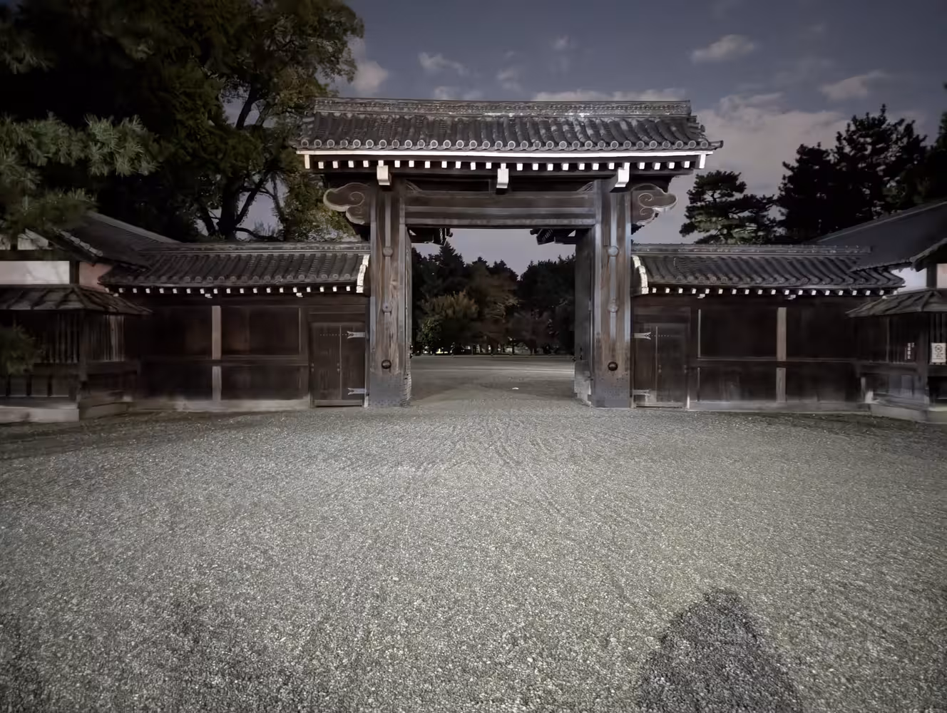 And open, old looking wooden gate at night with a large park on the other side.