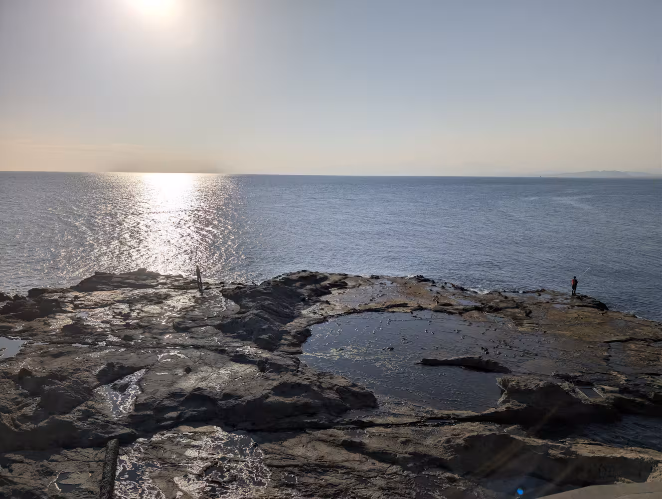 rocky cliffs in front of the blue ocean with the sun’s reflection. Two people are fishing on the rocks.