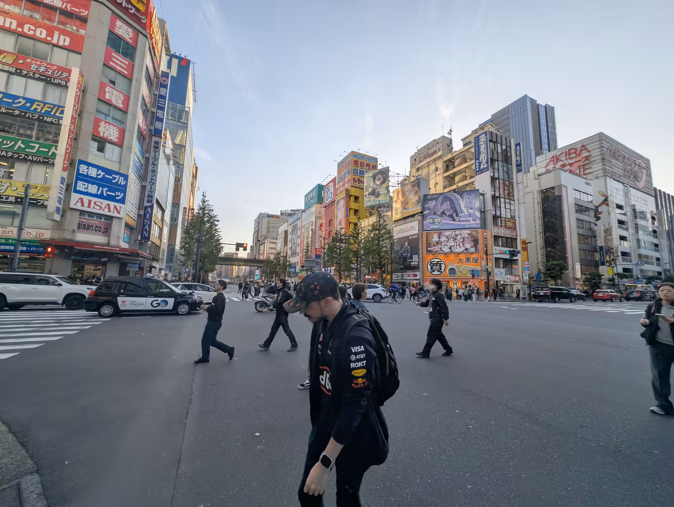 A large crossing in Akihabara. It is surrounded by mid-rise buildings with large billboards advertizing anime and similar things.