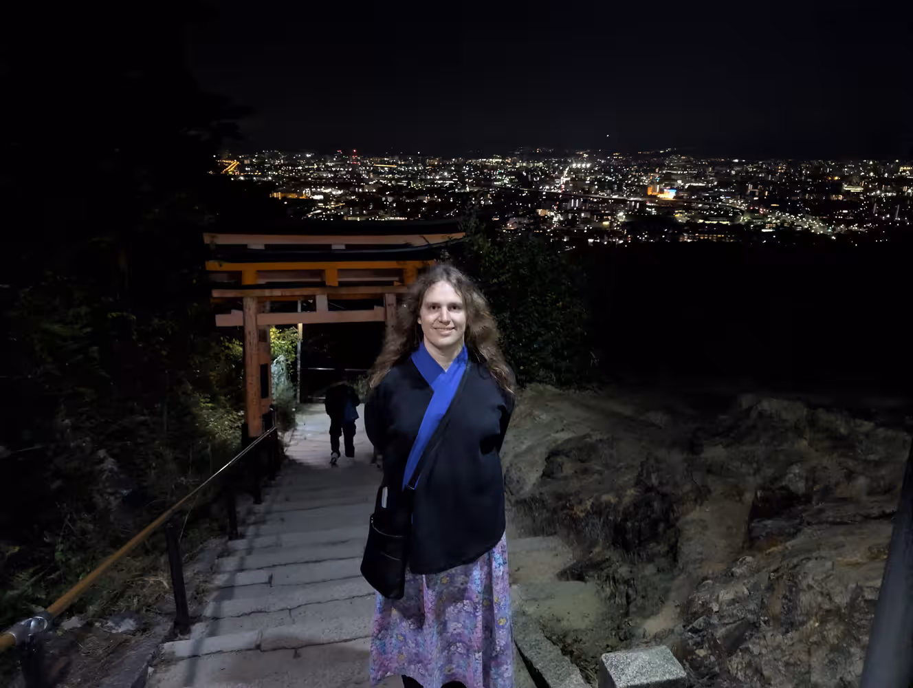 Fiona in front on the path downwards with some Torii visible directly behind her and the glowing city in the background.