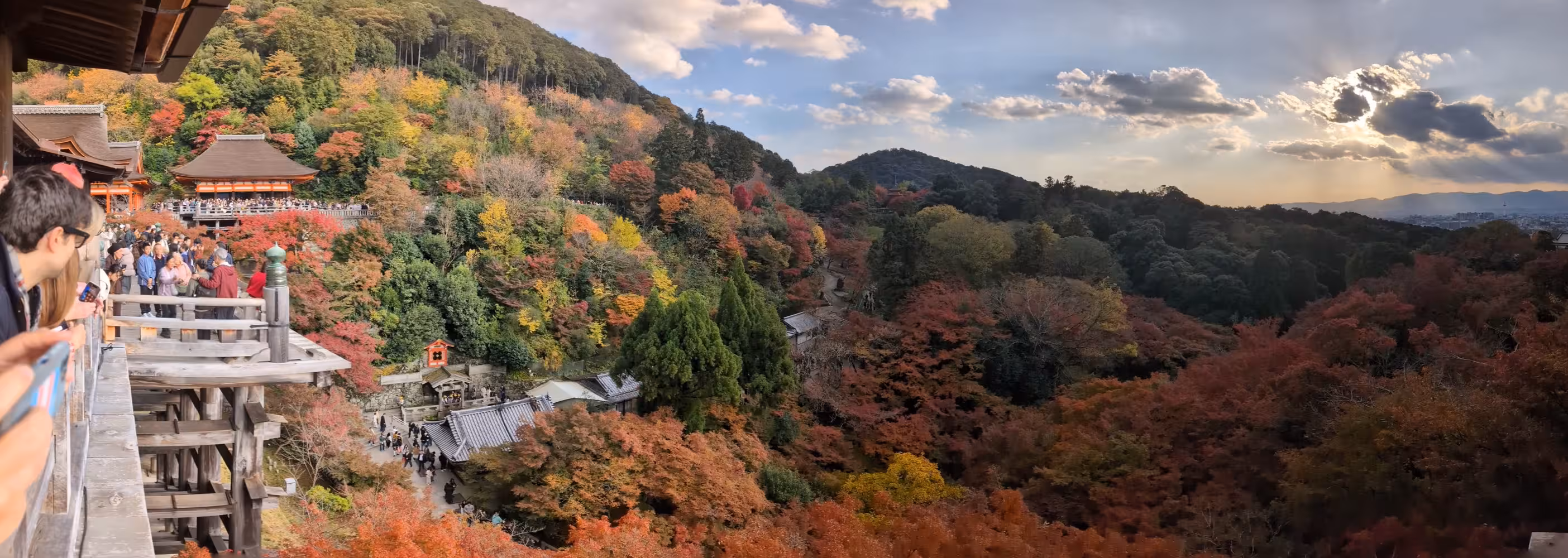 A panorama showing parts of the temple on the left, the valley with the colorful trees in the center and parts of Kyoto under the sun behind clouds.